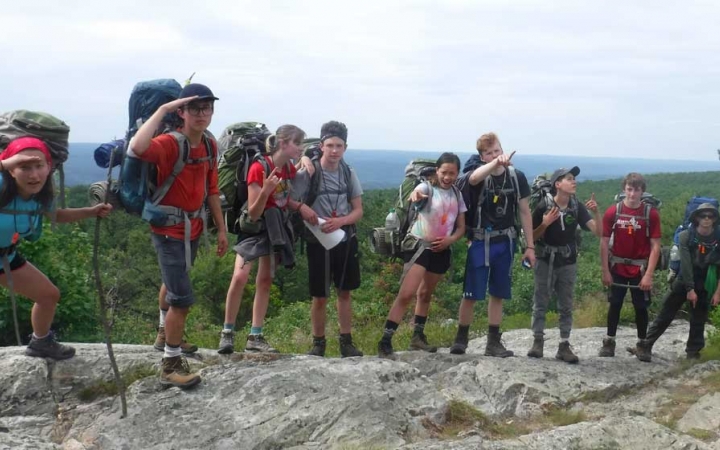 A group of young people wearing backpacks pose for a photo on a rock ledge with a vast green landscape behind them. 